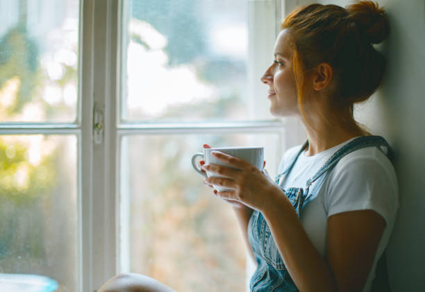 giovane bella donna sta guardando attraverso la finestra e bevendo caffè al mattino - guardare fuori foto e immagini stock