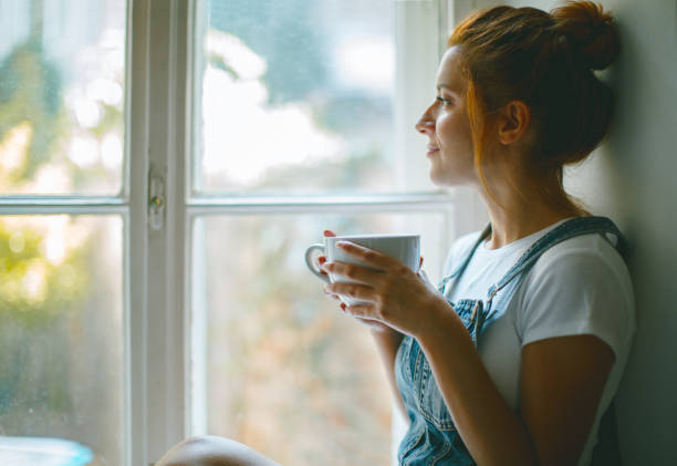 young beautiful woman is looking through the window and drinking coffee in the morning - coffee at home imagens e fotografias de stock