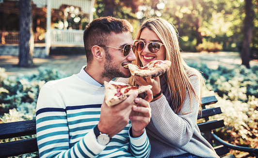 Beautiful young couple sitting in the park and eating pizza. Dating, consumerism, food, lifestyle concept