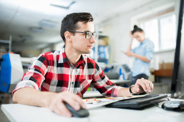 Young printing specialist working with computer Serious young printing specialist in glasses sitting at computer and working on design project in office digital printing house stock pictures, royalty-free photos & images