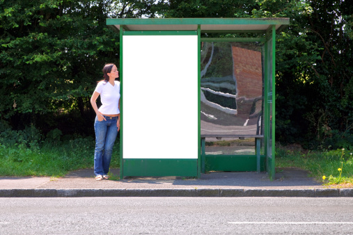 A woman standing at a rural bus stop leaning on a shelter with a blank billboard, clipping path included for you to add your own message in the space.