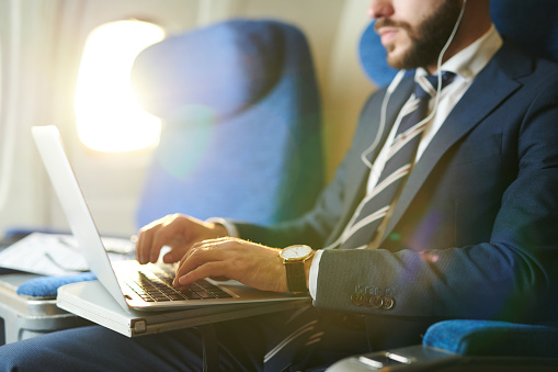 Mid section portrait of unrecognizable businessman typing on keyboard while using laptop during first class flight in plane, copy space