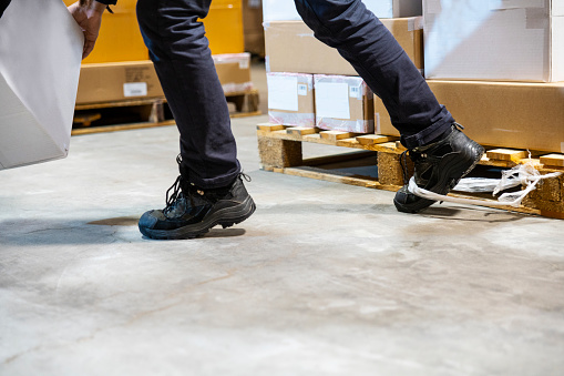 A warehouse worker tripping and falling beside a pallet.  His boot is caught in some plastic wrapping.