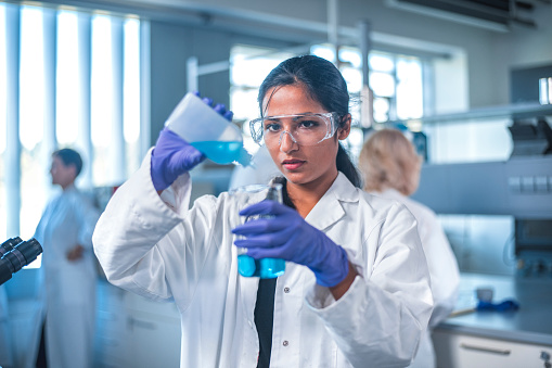 Young female researcher pouring chemical into flask. Confident scientist is doing experiment in laboratory. She is wearing protective eyewear.