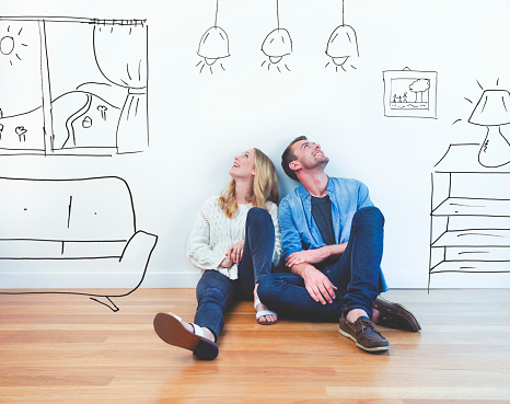 Couple dreaming of their new house. They are sitting on a wooden floor imagining their new home with furniture in it. The house is currently empty. They are booth looking up and smiling happily.