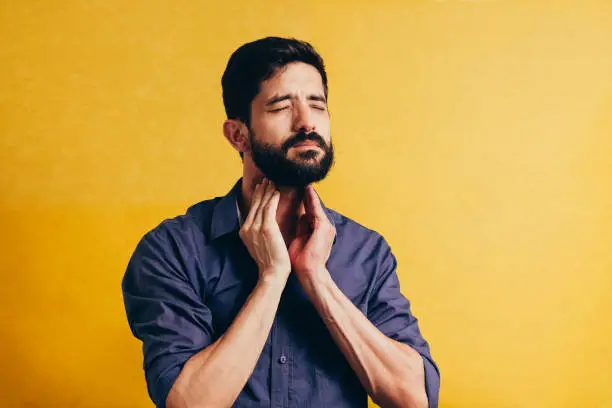 Photo of Young man having sore throat and touching his neck over yellow background. Hard to swallow