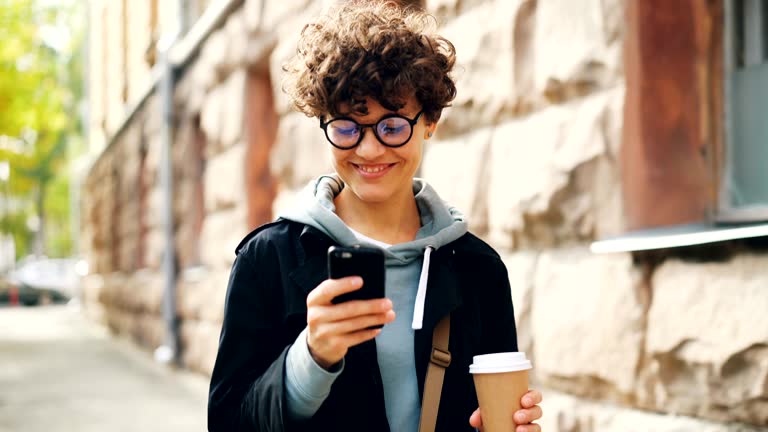 Smiling young woman in glasses is using smartphone looking at screen while walking outdoors in city with to-go coffee. Youth lifestyle, street and technology concept.