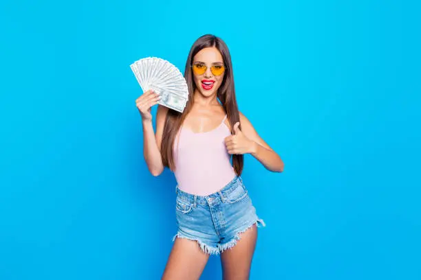 Nice confident cocky attractive adorable straight-haired girl wearing yellow sunglasses, showing fan of 100 usd, thumb-up, isolated over bright vivid blue background