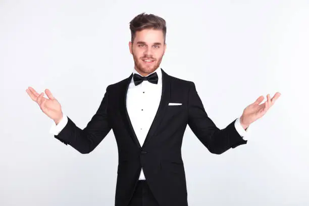 elegant man in tuxedo making a welcoming gesture while standing on light grey background