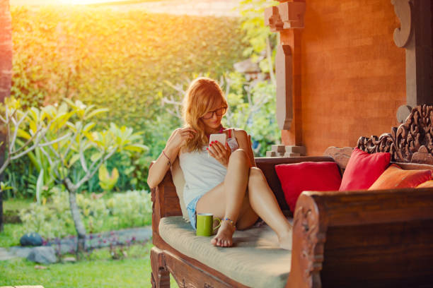 istock Young woman using cellphone and drinking coffee / tea on a porch sofa. 1048093322