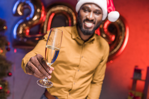 happy african american businessman in santa hat showing glass of champagne at new year corporate party