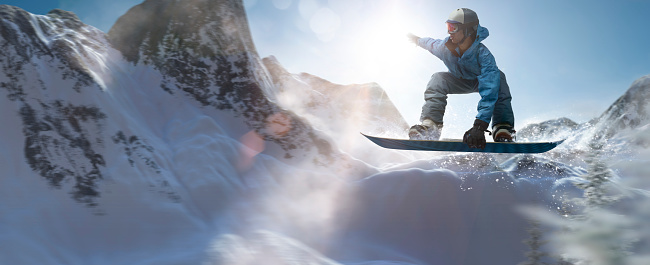 Panorama of a black male snowboarder in mid air during a jump close some trees on a snow covered mountain range. The snowboarder is wearing a generic blue salopettes, jacket, goggles and helmet and is squatting on snowboard with one hand held up. The action occurs in bright sunny daytime.