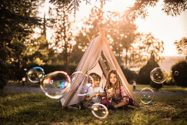 Two small siblings sitting in a tent in the backyard and playing with bubble wand.