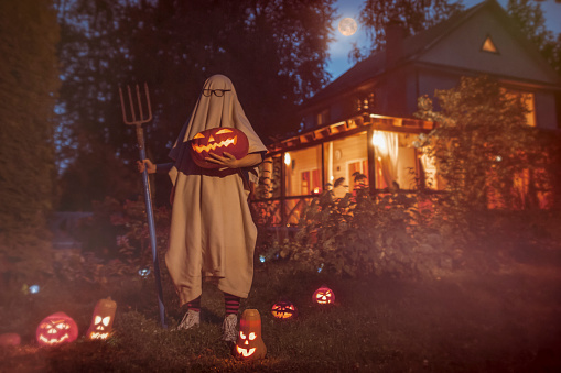 Child dressed as ghost standing near house in autumn at Halloween