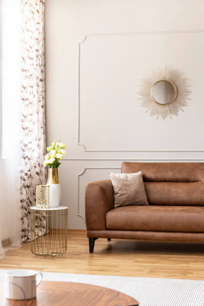 istock Stylish table with vase with flowers on it next to comfortable leather couch with beige pillow in luxury living room interior, real photo 1048216330