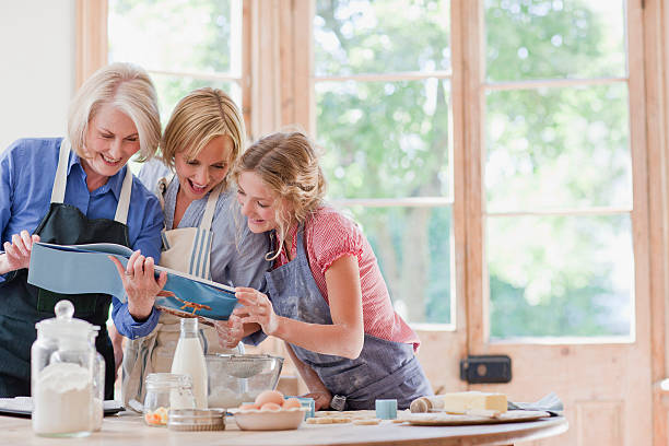 multi-generation frauen suchen kochbuch und beim backen in der küche, - looks back stock-fotos und bilder