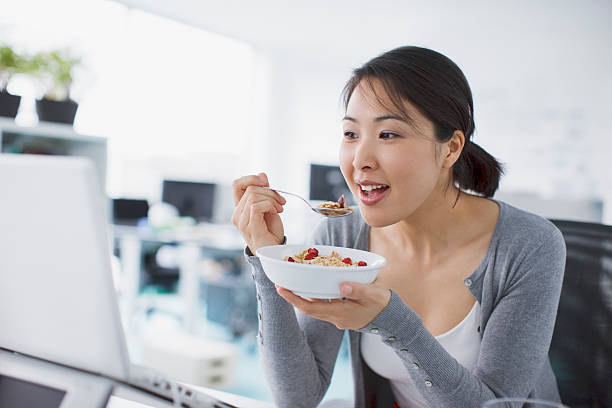 mulher de negócios comer cereais e olhando para laptop no escritório - eating women breakfast cereal imagens e fotografias de stock