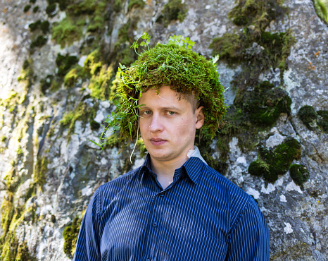 Office worker in a business suit, outdoors with a make-up on his face and green moss on his head. Man merges with nature