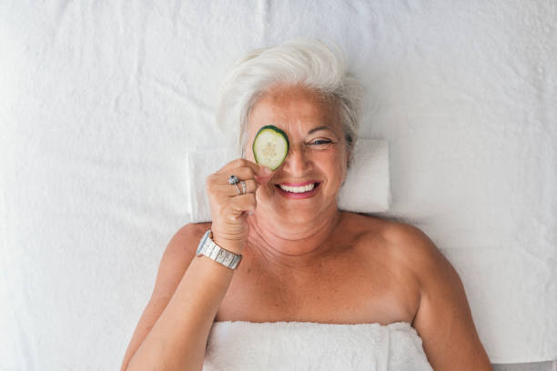 Photo of beautiful senior, gray hair, woman getting a facial at the spa with slice of cucumber in front of her eye. Portrait of woman with cucumber treatment on her face have a relax in a wellness center. Concept of skincare, cosmetics, beauty salon, facial treatment, facial massage, wellbeing