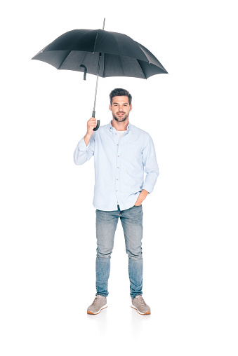 full length view of handsome young man holding black umbrella and looking at camera isolated on white