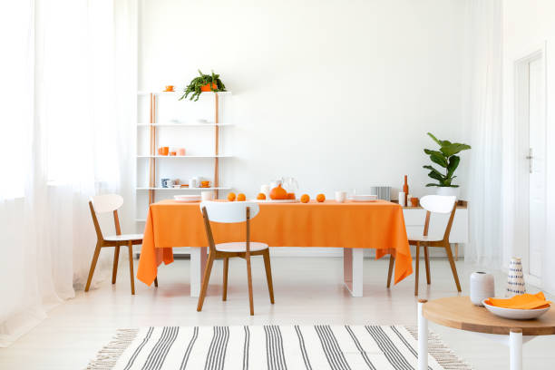 istock Long dining room table covered with orange tablecloth and comfortable white chairs around it. 1048260192