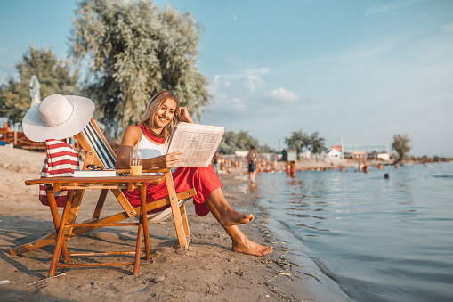 Portrait of attractive young woman reading newspaper while sitting on deck chair on the beach