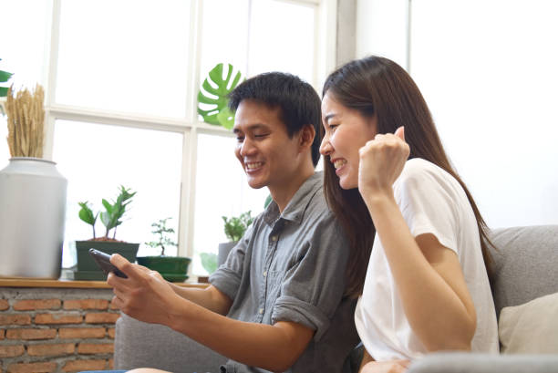 istock Young couple sitting on sofa are watching mobile phone and feeling surprise&happy when know the result with smile face. Happy family relationship concept. 1048273886