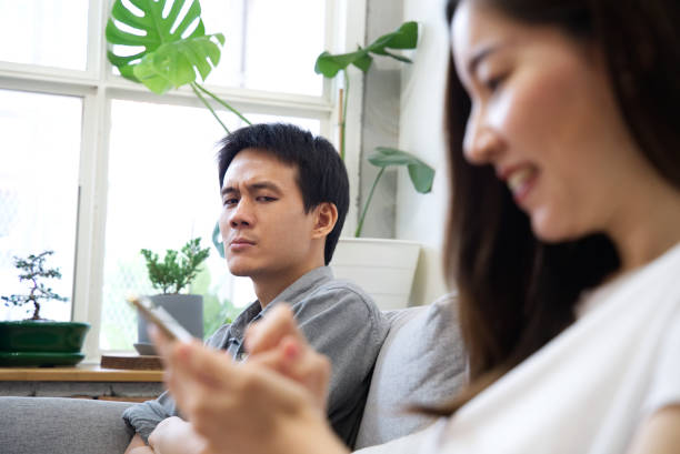 A Man sitting on sofa is feeling unhappy with his girlfriend while she enjoy using the phone and ignoring him. She spent too much time on the phone. Social problem in family relationship concept. Bad behavior from social problem asian boyfriend too much phone stock pictures, royalty-free photos & images