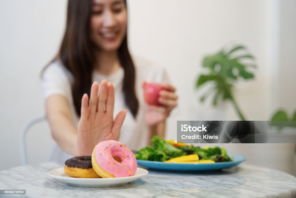 Young girl on dieting for good health concept. Close up female using hand reject junk food by pushing out her favorite donuts and choose red apple and salad for good health. Healthy food concept. Food Stock Photo