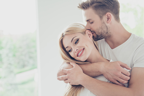 Portrait of beautiful attractive sweet lovely charming cute nice adorable couple, spouses, husband and wife in white t-shirts on honey-moon at hotel, embracing in light interior room