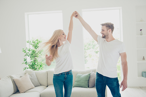 Beautiful attractive charming cute nice adorable playful cheerful couple in white t-shirts and jeans in house, holding hands, dancing, celebrating holiday in light interior room