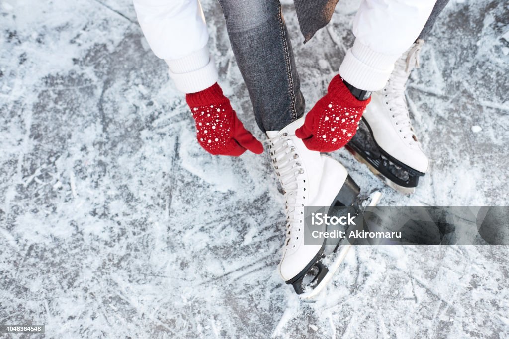 Girl tying shoelaces on ice skates before skating on the ice rink, hands in red knitted gloves. Girl tying shoelaces on ice skates before skating on the ice rink, hands in red knitted gloves. View from top. Ice-skating Stock Photo