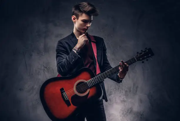Photo of Handsome young thoughtful musician with stylish hair in elegant clothes posing with a guitar in his hands.