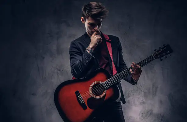 Photo of Handsome young thoughtful musician with stylish hair in elegant clothes posing with a guitar in his hands.