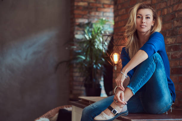 istock Beautiful blonde female sitting on a table against a brick wall in a studio with a loft interior. 1048401106