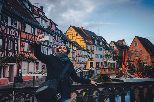 Lovely young tourist male enjoying Christmas, holding smart phone and taking selfie pictures. He is happy, smiling enjoying and looking at camera against water canal and traditional colored houses reflected in river Lauch at Noel time in Little Venise Colmar Haut-Rhin department Alsace France Europe