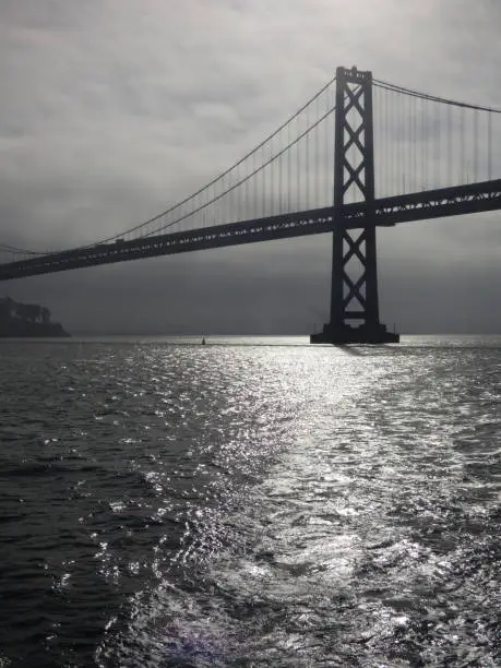 SF Bay Bridge seen from the water with wake of boat. Reflection of sunrise and dark sky