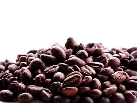 Close up of a isolated dense pile or heap of aromatic roasted brown coffee beans on a high key white minimalist background with soft natural morning sunlight reflecting on it