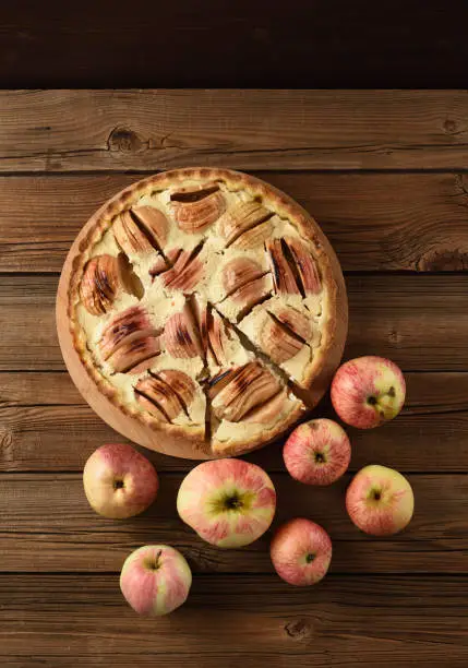 Homemade apple pie with cream filling served with raw organic apples on old wooden background overhead view
