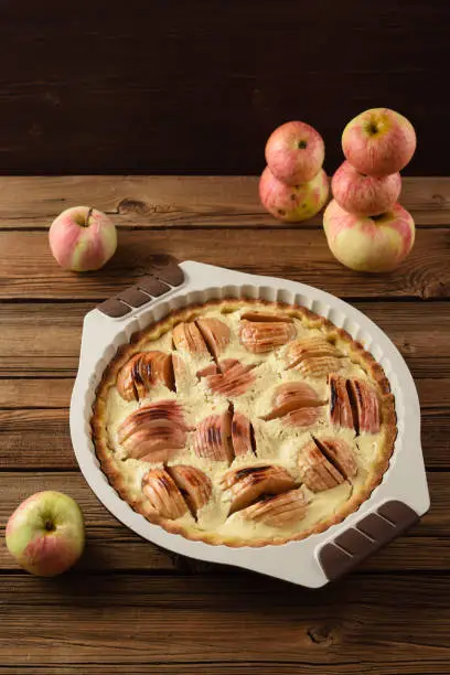 Tasty freshly baked apple tart with cream filling in baking dish served with organic apples on old wooden background copyspace