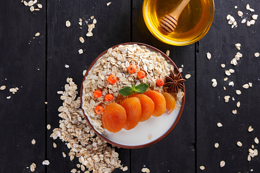 Oatmeal with dried apricots and honey on black background. Oat flakes with milk and green mint on dark wooden boards. Healthy food for vegans. Dry oats with berries of mountain ash. Autumn pattern