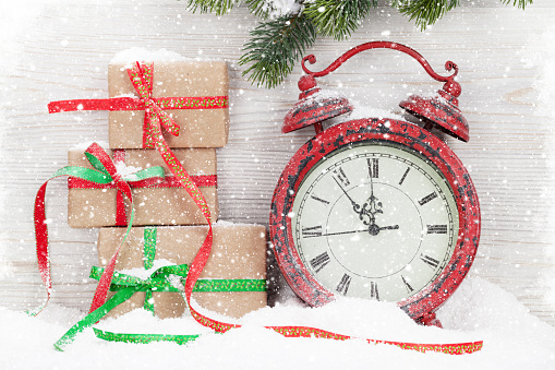 Christmas gift box, alarm clock and fir tree branch covered by snow in front of wooden wall