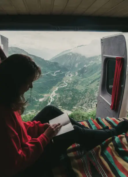 Young Caucasian woman reading book in camper van with view on Furka Pass