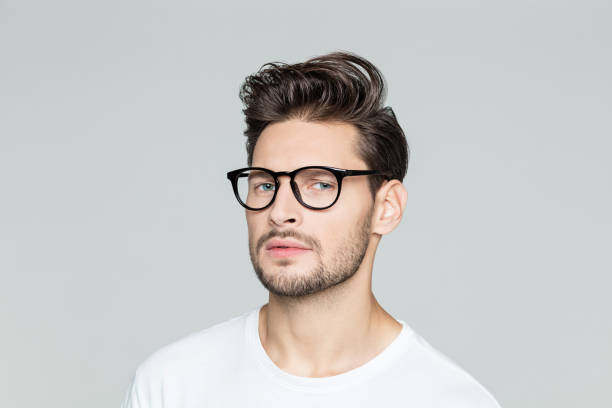Close up portrait of young man with beard wearing eyeglasses looking at camera against grey background