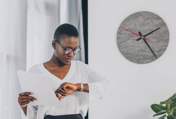 stylowa afroamerykańska bizneswoman trzymająca dokumenty i sprawdzająca czas w biurze, zegar na białej ścianie - checking the time time clock women zdjęcia i obrazy z banku zdjęć