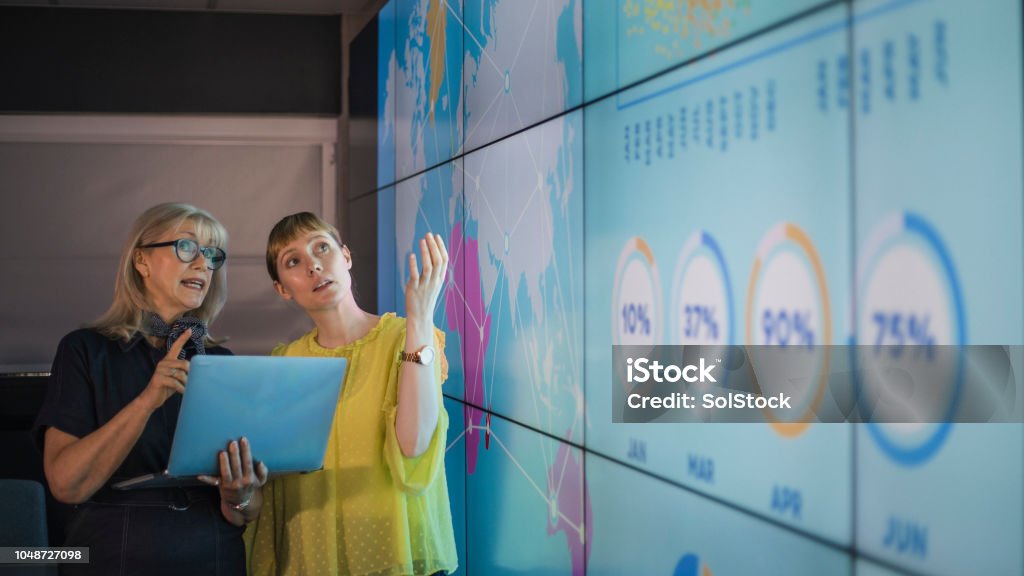 Businesswomen Discussing Ideas Against an Information Wall An experienced woman mentors a female colleague, the mature woman is holding a laptop as they debate data from an interactive display; they are both wearing smart casual clothing. Data Stock Photo