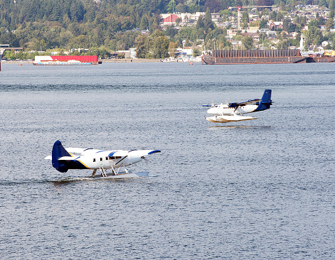 Two twin engine light aircraft seaplanes passing on the sea at Vancouver Harbour, one touching down to land whilst the other, floating past is taxiing to take off, Vancouver, B.C., Canada