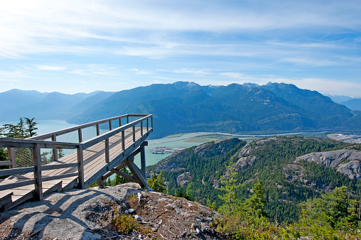 Aerial walkway overlooking the mountain, coniferous forest and woodland - an observation platform that extends over the cliff precipice, with peaks under the dramatic wispy cloudscape in the distance, near Squamish, Vancouver, B.C., Canada