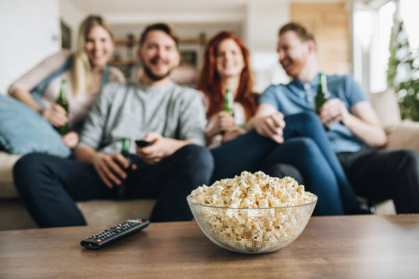 Close up of popcorns in a bowl with friends in the background. Close up of popcorn in a bowl and remote controller on the table with friends in the background. remote control on table stock pictures, royalty-free photos & images