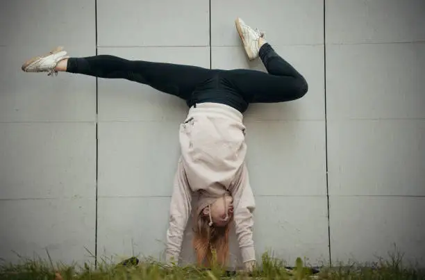 Acrobat girl in a beige hoodie performs the splits while standing on hands on a background of the wall. Wide shot